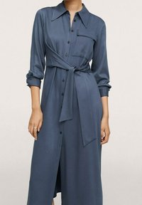 Abito camicia blu con vita legata, maniche lunghe, chiusura con bottoni e tasca sul petto. Realizzato in tessuto morbido con una leggera lucentezza.