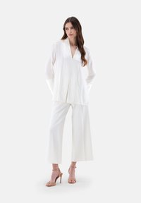 Weiße Satinbluse mit tiefem V-Ausschnitt und breiten Ärmeln, kombiniert mit weißen weitem Hosen. Sandalen mit Absatz runden das Outfit ab.