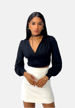 Top lungo nero a maniche coordinate con scollo a V profondo e dettagli arricciati, abbinato a una mini gonna bianca. Orecchini a cerchio dorati e un braccialetto completano il look.