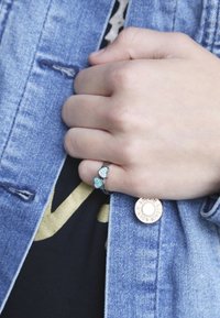 Zilveren ring met twee hartvormige turquoise stenen, gedragen aan een hand die een spijkerjack vasthoudt, naast een ronde metalen knoop.