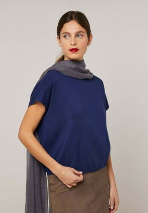 Femme aux cheveux bruns portant un haut bleu à manches courtes, une jupe marron, une écharpe grise drapée sur les épaules, et un rouge à lèvres rouge, regardant sur le côté.