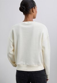Sudadera de color crema con un corte relajado, cuello redondo y puños acanalados. Parece estar hecha de un tejido suave y texturizado. Vista trasera sin adornos.