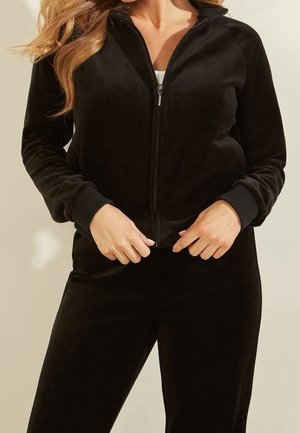 Veste noire à fermeture éclair en tissu doux et velouté. Comprend des poignets côtelés et une texture lisse, associée à un pantalon assorti.