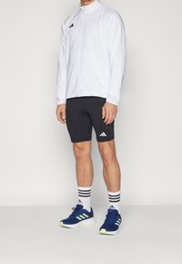 Veste de sport blanche à col montant, logo noir ; shorts noirs ; baskets bleues avec accents verts et chaussettes noires à rayures.