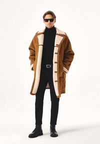 Pruun shearling- mantel kreemjast voodriga, millel on toggli kinnitused, kantud musta kõrge kaelusega särgi peal, koos mustade pükste ja nööpsärkide kingadega.