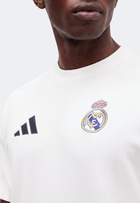 Homme portant un maillot de sport blanc avec le logo du Real Madrid et un emblème Adidas noir sur un fond clair uni.