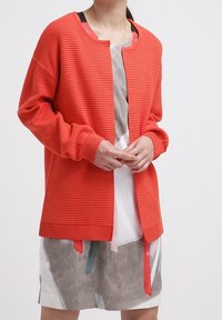 Orange ribbad kofta med rund halsringning, långa ärmar och knäppning fram, bärs över en lagrad klänning med abstrakta mönster och band.