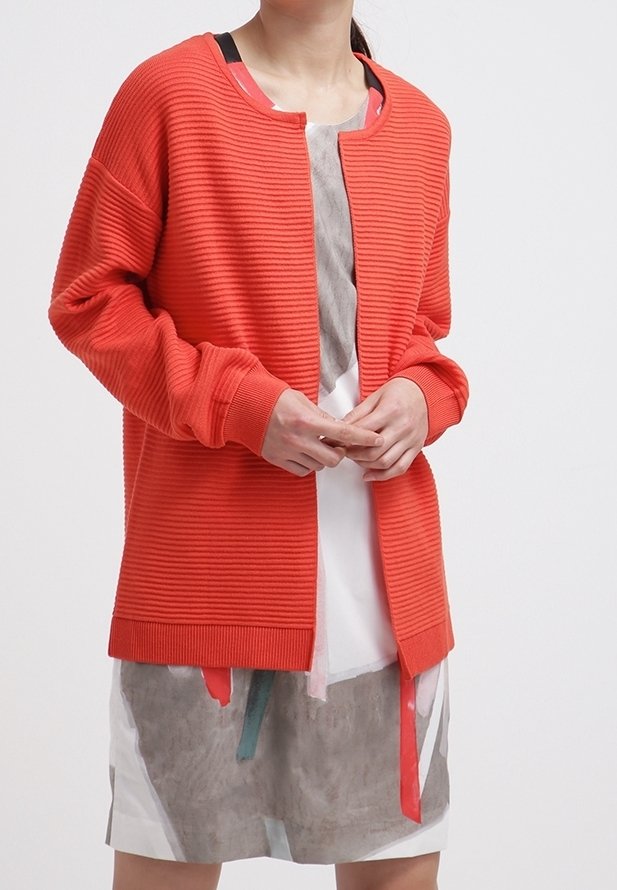 Orange ribbad kofta med rund halsringning, långa ärmar och knäppning fram, bärs över en lagrad klänning med abstrakta mönster och band.