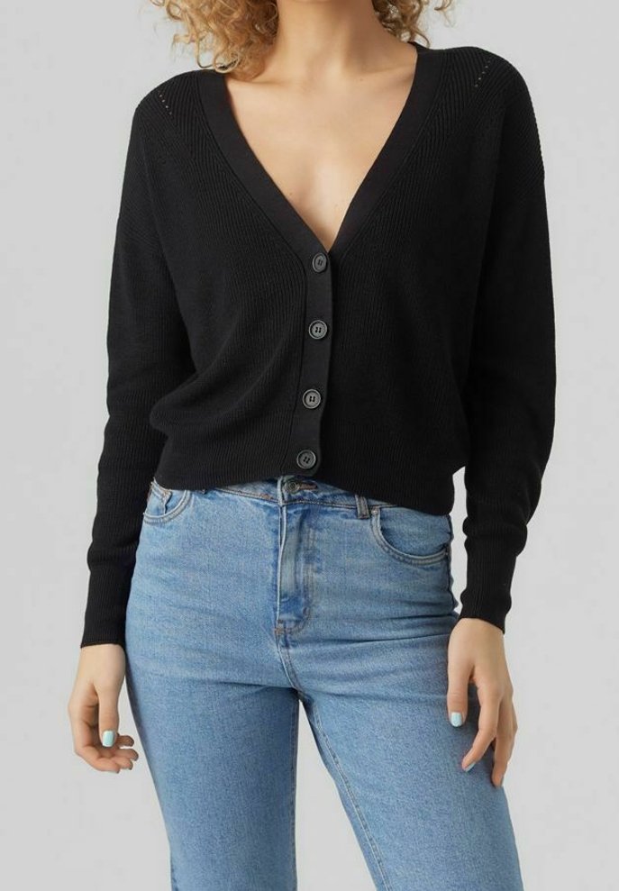 Femme portant un cardigan noir à col en V boutonné et un jean bleu clair taille haute, se tenant devant un fond gris uni.