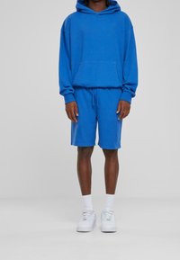 Sudadera de algodón azul con bolsillo delantero y cordón, combinada con pantalones cortos azules a juego. Se lleva con zapatillas blancas y calcetines hasta la altura del tobillo.