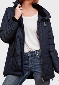 Chaqueta aislante azul marino con cierre frontal de cremallera, botones a presión y cuello forrado de pelo sintético. Incluye cordones ajustables en el dobladillo.