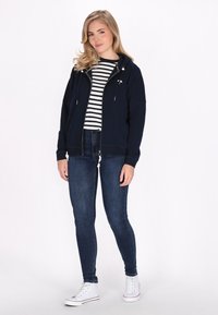 Felpa con zip navy e cappuccio con cordini sopra una maglietta a righe bianche e nere, abbinata a jeans skinny scuri e sneakers bianche.