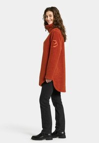 Kauranvärinen, pehmeä fleece-pusero, jossa on korkea kaulus, sivuhalkiot ja merkki vasemmassa hihassa; yhdistetty mustiin slim-fit-farkkuihin ja saappaisiin.