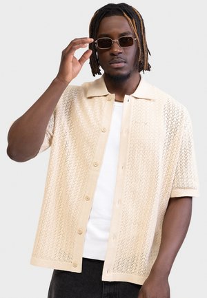 Άνδρας με μέτρια σκουρόχρωμο δέρμα και ντραντλoκς ρυθμίζει ορθογώνια γυαλιά ηλίου, φορώντας μία μπεζ κοντομάνικη πουκαμίσα ανοιχτού πλεξίματος πάνω από ένα λευκό T-shirt.