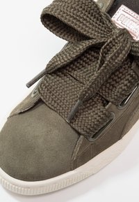 Gros plan sur une basket en daim vert olive avec des lacets tressés épais noués en nœud et une semelle en caoutchouc blanche.