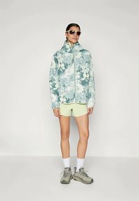 Bunda s kvetinovým vzorom v tlmených modrozelených odtieňoch, so zipsom na prednej strane, skombinovaná s svetlozelenými šortkami a šedými teniskami.