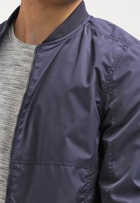 Giubbotto bomber blu con una texture liscia e lucida e colletto a coste, abbinato a una camicia a righe grigio chiaro sotto.