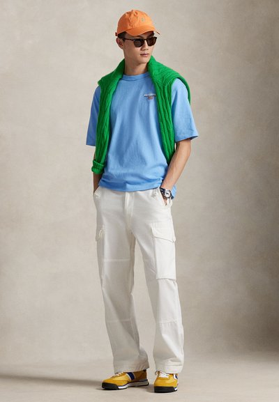 Καθημερινή ενδυμασία που περιλαμβάνει ένα ανοιχτό μπλε t-shirt, ένα πράσινο πλεκτό πουλόβερ δεμένο γύρω από το λαιμό, λευκά παντελόνια cargo και κίτρινα αθλητικά παπούτσια με μπλε λεπτομέρειες.