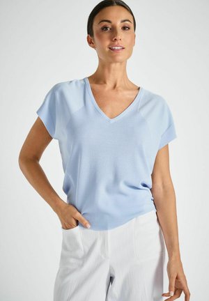 Femme portant un haut bleu clair à manches courtes et col en V, et un pantalon blanc, posant avec une main dans la poche devant un fond uni.