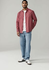 Chemise à boutons à motifs rouges portée par-dessus un t-shirt blanc, associée à un jean bleu clair et des baskets blanches à rayures noires.