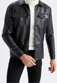 Veste en cuir noir avec deux poches poitrine, des boutons à l'avant et une texture lisse, portée sur un pull en maille blanche. Design ajusté.