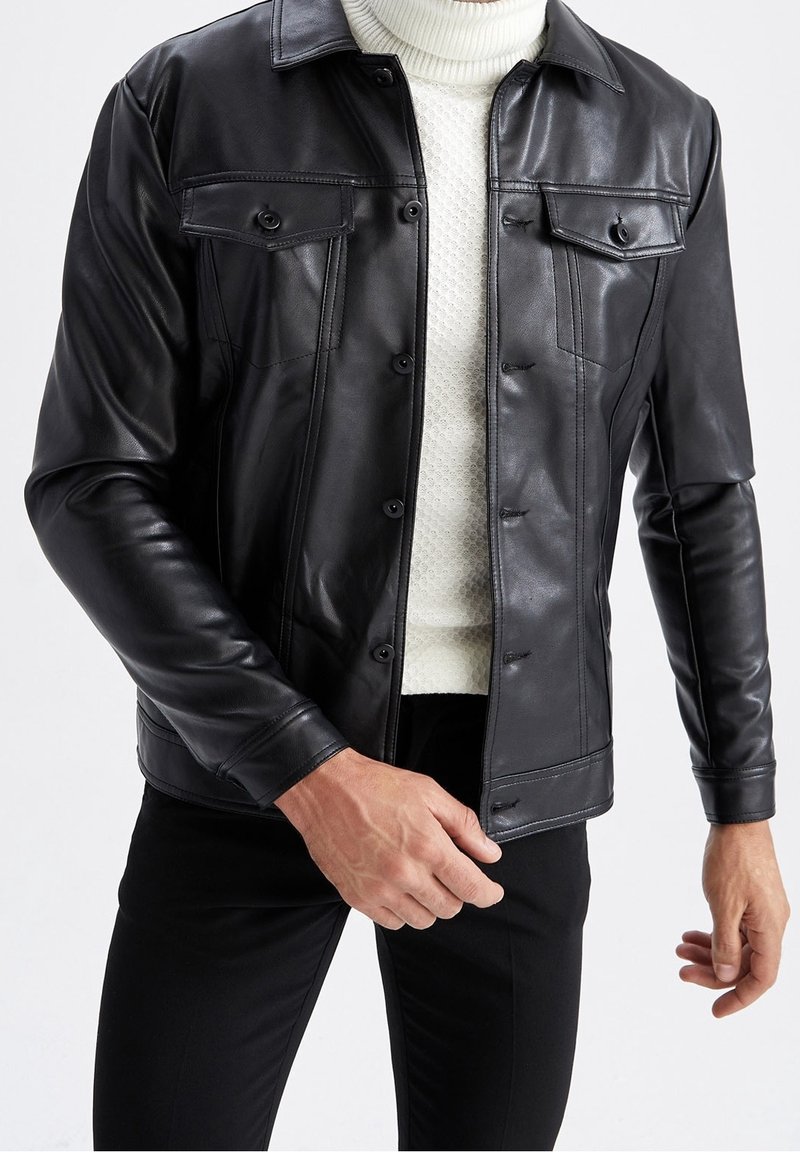 Veste en cuir noir avec deux poches poitrine, des boutons à l'avant et une texture lisse, portée sur un pull en maille blanche. Design ajusté.