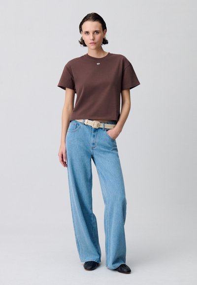 T-shirt marron à manches courtes avec un col rond ; associé à un jean bleu clair à jambes larges et une ceinture tressée beige. Des chaussures noires complètent le look.