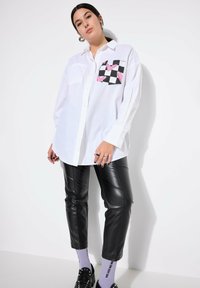 Camicia bianca in cotone con bottoni e taschino a scacchi che presenta cuori rosa; abbinata a pantaloni in pelle neri e calzini viola.