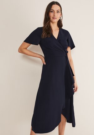 Vrouw met lang bruin haar die een donkerblauwe wikkeljurk met korte mouwen draagt, met één hand op haar heup staand tegen een effen achtergrond.