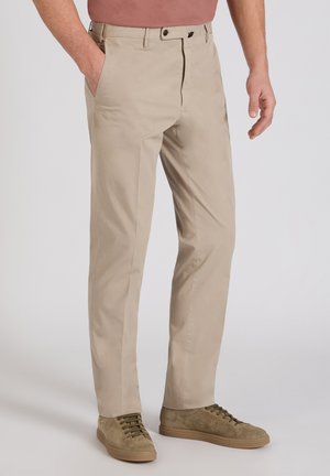 Pantaloni beige casual a taglio dritto con bottone frontale e cerniera, indossati con sneakers in camoscio color oliva, mano in tasca e camicia color ruggine.