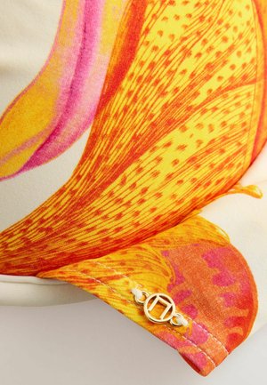 Lysegult og orange blomsterstof med lyserøde accenter, med en lille rund guldbesked fastgjort med hvid tråd på stoffets kant.