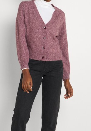 Femme portant un cardigan mauve boutonné sur un pull à col roulé blanc et un pantalon noir, debout devant un fond uni.