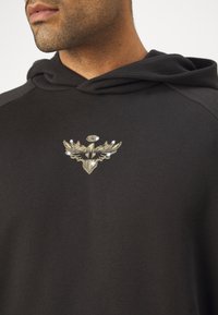 Hanorac negru cu o textură moale, prezentând un emblema metalică aurie cu aripi și un accent circular pe piept.