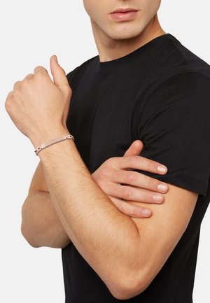 Metal bracelet in soft pink with a streamlined design, featuring decorative links. Worn on a man's wrist, paired with a black t-shirt.
