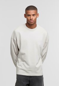 Camiseta de manga larga gris claro con cuello redondo, corte relajado y textura suave. El modelo está de pie con las manos detrás de la espalda frente a un fondo liso.