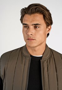 Olivgrön bomberjacka med ribbad krage, med en vadderad design. Bärs över en svart ribbad tröja. Neutral bakgrund.