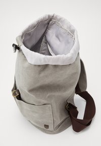 Mochila de lona gris claro con interior de tela a rayas, bolsillo frontal, herrajes en tono dorado y correa de hombro ajustable de color marrón oscuro.
