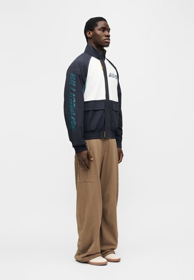 Άνδρας που στέκεται πλευρικά, φοράει μια ναυτική, λευκή και τιρκουάζ ζακέτα με φερμουάρ και κείμενο, μπεζ παντελόνι με φαρδιά σκέλη και λευκά παπούτσια σε απλό φόντο.