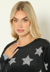 Svart långärmad topp med vita stjärnmönster, slät textur och rund halsringning. Anmärkningsvärd detalj: stjärndekorationer på ärmarna.