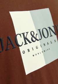 Brun bomulds t-shirt med et rektangulært grafisk design i hvide og grå paneler; fed sort tekst står der "JACK & JONES ORIGINALS WORLDWIDE."