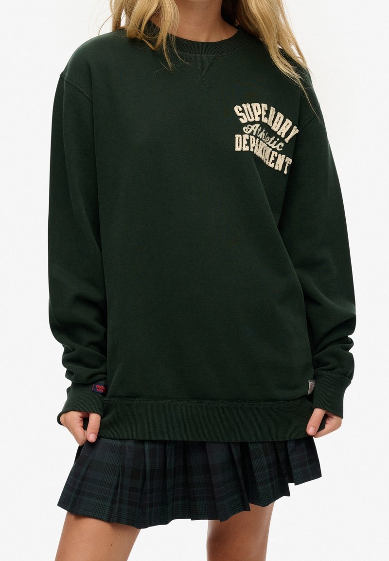 Personne portant un sweat-shirt Superdry vert foncé avec un texte brodé sur la poitrine et une jupe plissée à carreaux foncée.