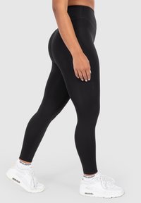 Person trägt schwarze hochgeschnittene Leggings und weiße Sneakers mit gebrandeten Socken, steht seitlich auf einem einfarbigen hellen Hintergrund.