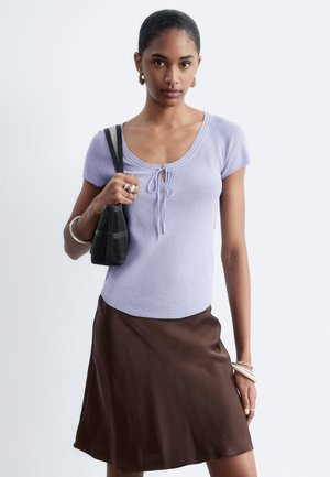 Femme portant un haut violet clair à manches courtes et une jupe marron, tenant un sac à main noir sur son épaule devant un fond uni.