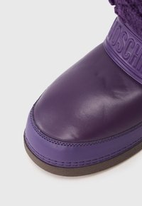 Botín de cuero morado con un acabado suave, que presenta una parte superior de tela morada texturizada y una marca en relieve en la correa. Diseño de punta redonda.