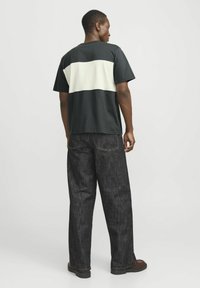 Camiseta negra con una franja horizontal crema en la parte posterior, combinada con jeans oscuros de corte holgado y zapatos negros. Diseño simple, estilo casual.