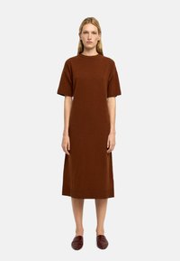 Brązowa dzianinowa sukienka z krótkimi rękawami, długości do kolan, o luźnym kroju, z okrągłym dekoltem i gładką strukturą. Zestawiona z ciemnoczerwonymi płaskimi butami.