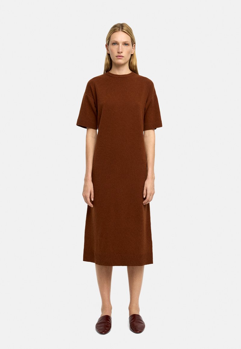 Brązowa dzianinowa sukienka z krótkimi rękawami, długości do kolan, o luźnym kroju, z okrągłym dekoltem i gładką strukturą. Zestawiona z ciemnoczerwonymi płaskimi butami.