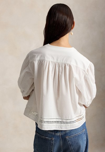 Γυναίκα με μακριά σκούρα μαλλιά που φοράει λευκό μπλούζα με φουσκωτά μανίκια και λεπτομέρεια κοψίματος στο κάτω μέρος, καθώς και μπλε τζιν, γυρισμένη με την πλάτη στην κάμερα.
