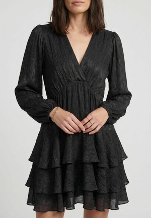Femme portant une robe noire texturée à manches longues avec une jupe à volants et un profond décolleté en V, mains jointes montrant des bracelets et des bagues en or.
