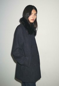 Sort dobbeltbrystet uldfrakke med faux pelskrave, med store knapper og lommer i siden, glat tekstur og afslappet pasform.